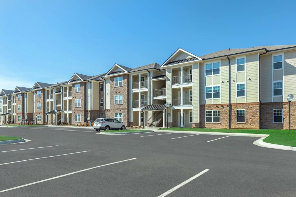 A line of brand new apartment complexes. The buildings are three stories tall. The first floor is made of brown stone and the others of a pale yellow/green siding. Only one car sits in the newly paved parking lot.