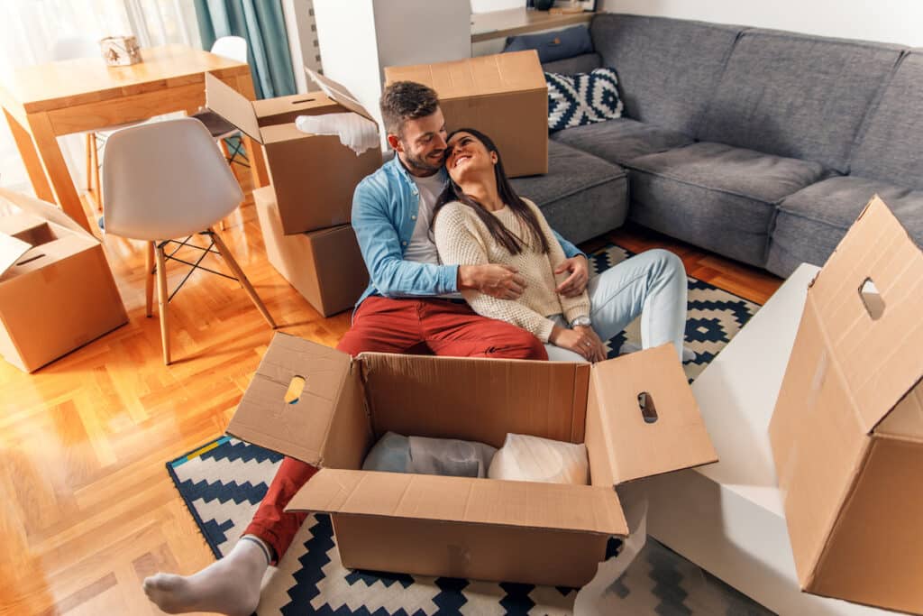 A smiling young couple lean against each other on the floor of a new apartment. A box with wrapped pieces of pottery sit in front of them and more cardboard boxes can be seen in the background on the sofa and beside a table.