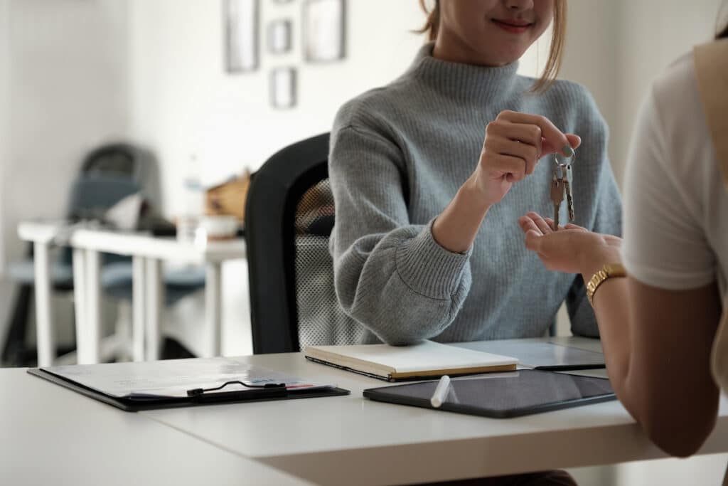 A real estate agent sitting behind her desk handing a set of two silver keys to a woman's hand wearing a gold watch. The desk has a clipboard, tablet, and a notepad sitting on it. Only the bottom half of the real estate agent's face can be seen.