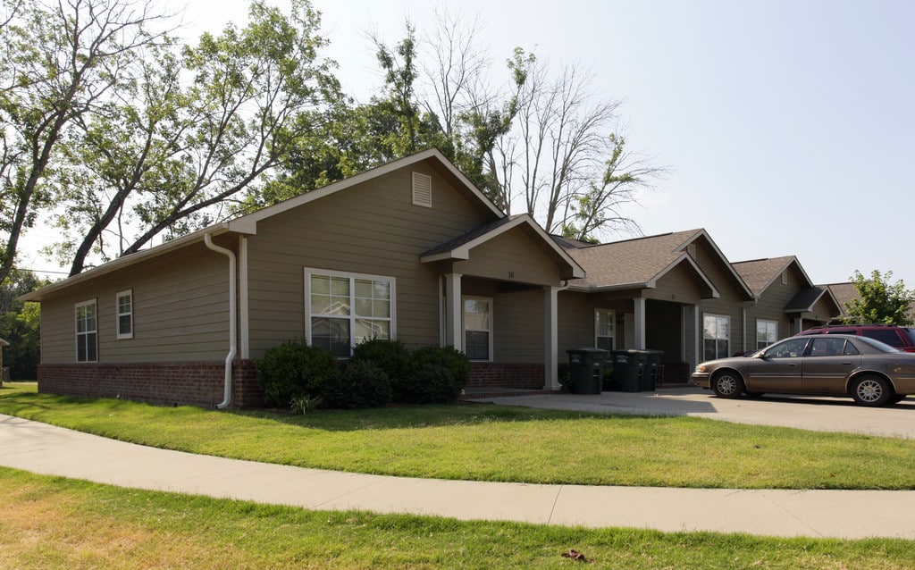Stonegate Town Homes, managed by Welcome Home Properties. The homes are a dark brownish green color in the shade and are all connected with a shared driveway. There is a small green lawn in front and bushes and trash cans sit in lines in front of the building.